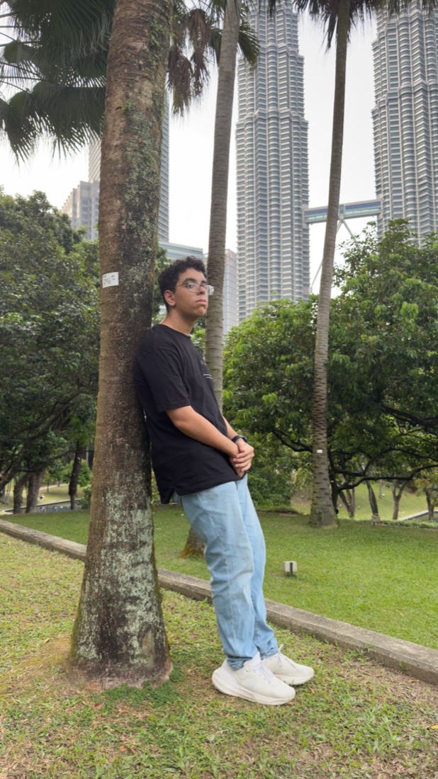 Yousof Selim leaning against a palm tree in KLCC Park, the Petronas Towers behind him